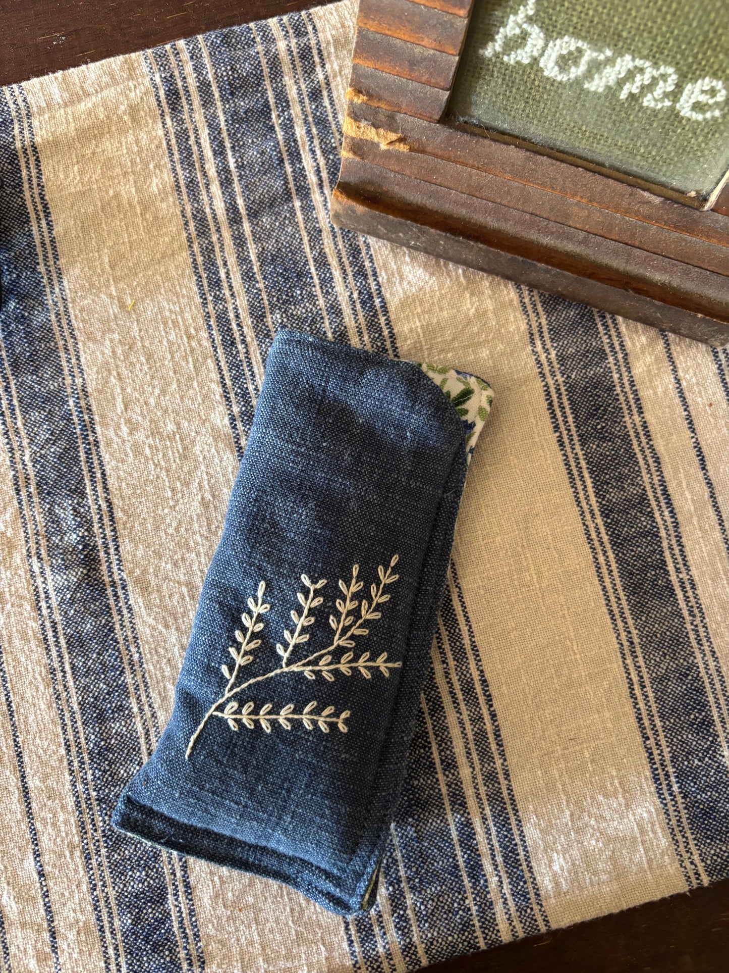 Blue embroidered napkin on a striped fabric surface with a wooden frame in the corner.