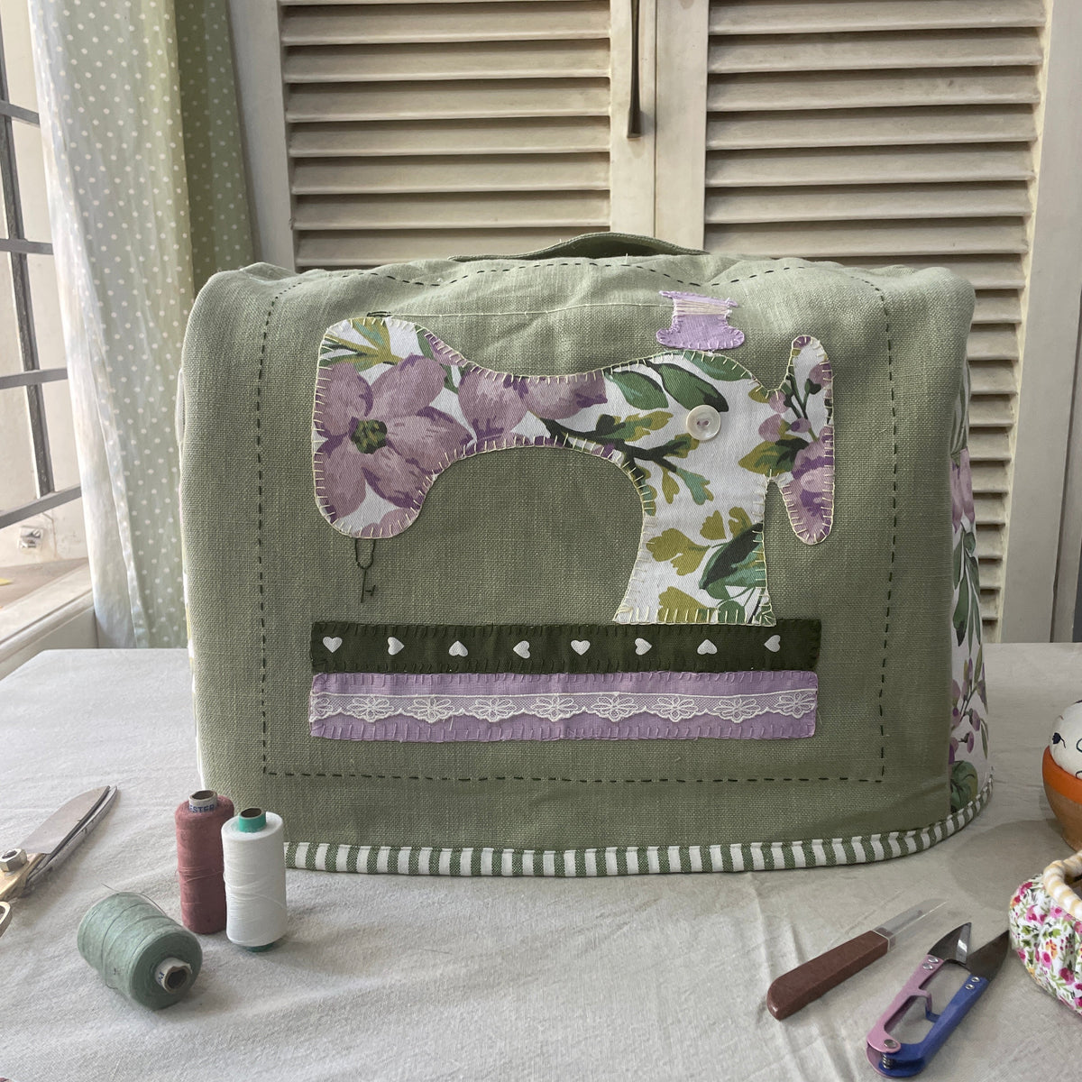 A green sewing machine cover with floral applique design, displayed on a sewing table with various sewing-related items around it.
