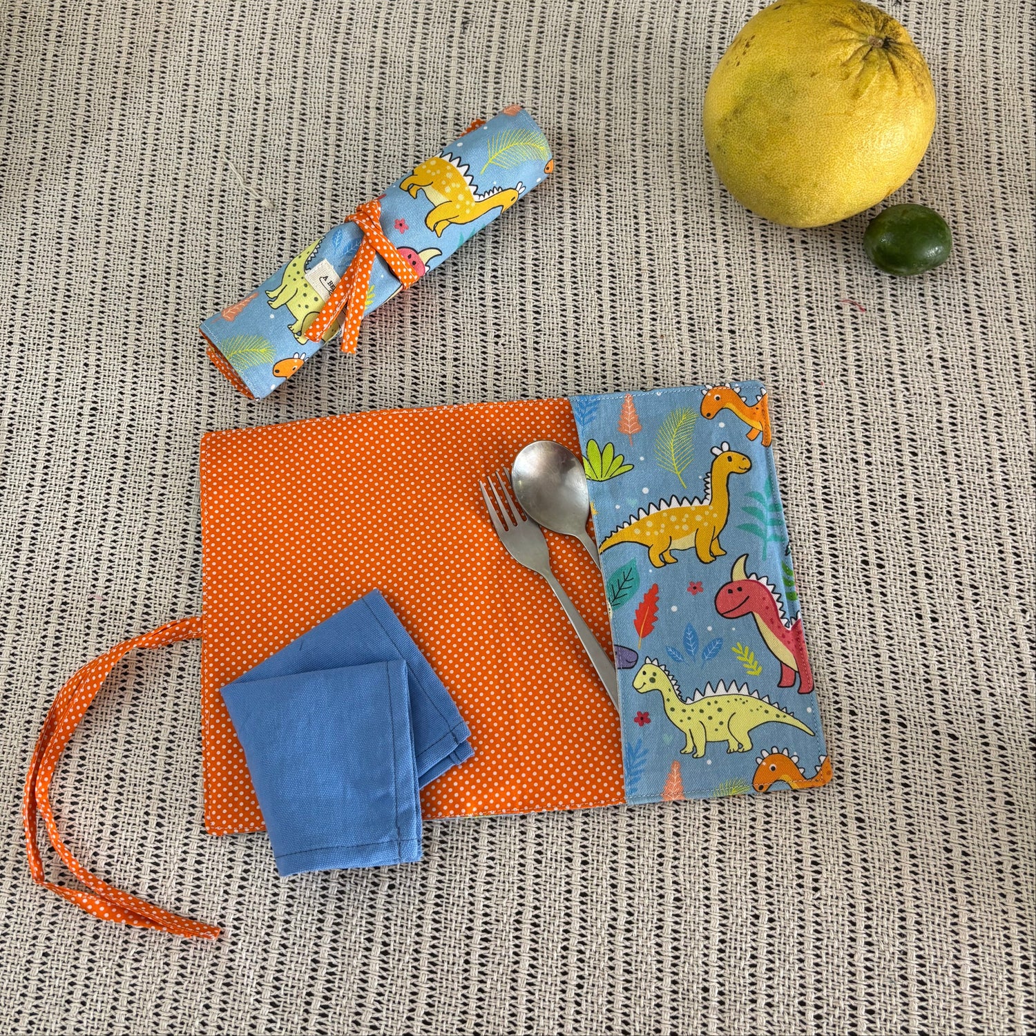 Children's tableware set with dinosaur-themed cutlery holder, napkin, and placemat on a textured surface.