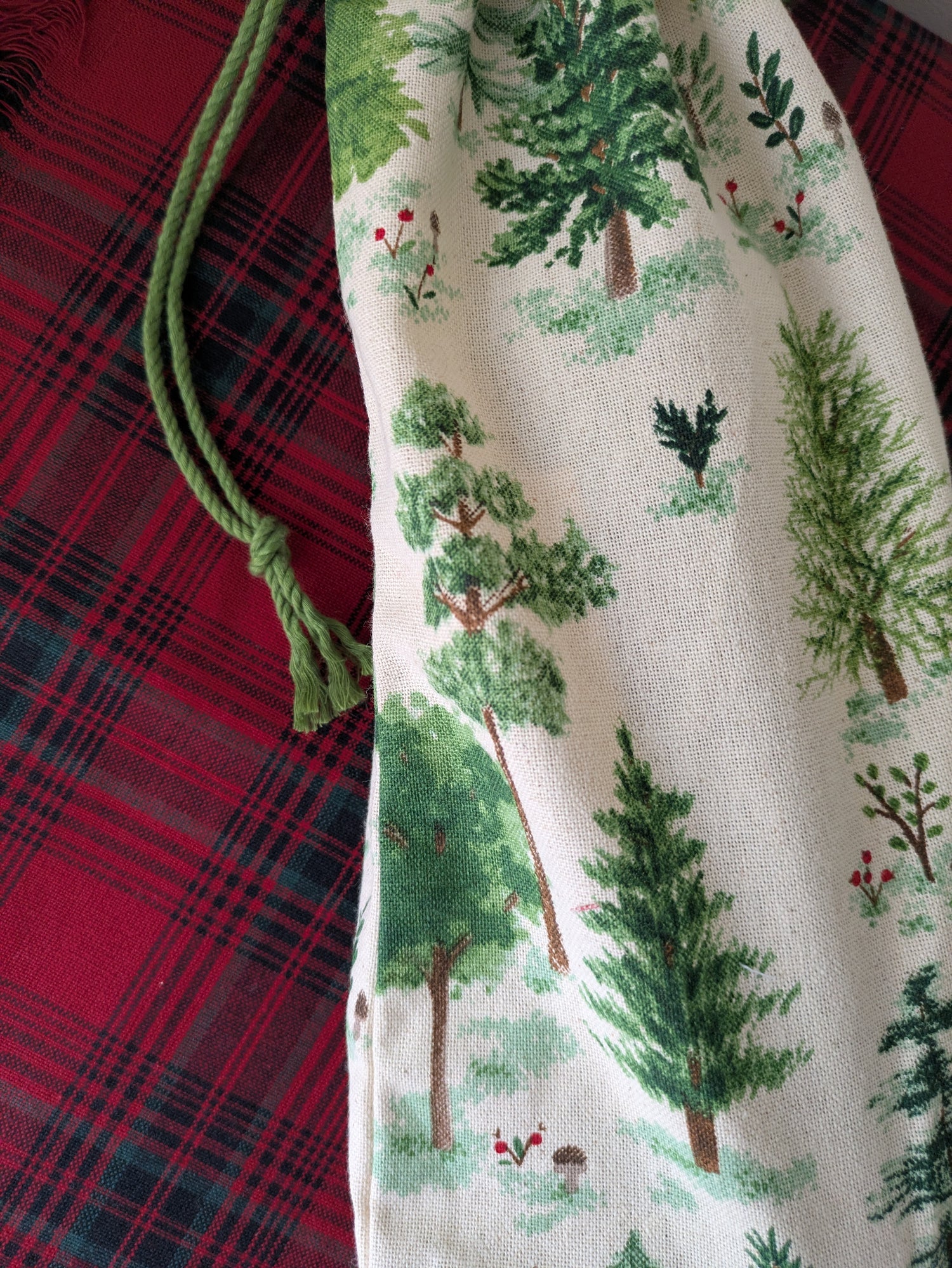 Fabric drawstring bag with green tree pattern held by a hand on a red plaid background