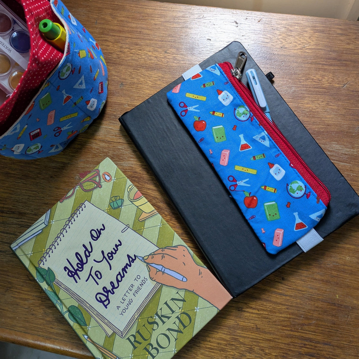 A blue pen sleeve with a pattern of books, pens, and other stationery items, displayed on top of a black hardbound journal or notepad.