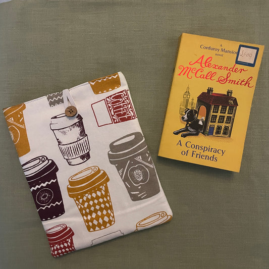 A book sleeve with a coffee cup pattern next to a book titled 'A Conspiracy of Friends' by Alexander McCall Smith.